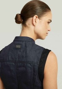 Gilet in denim blu scuro con design senza maniche, caratterizzato da una toppa con logo sul retro, colletto strutturato e texture liscia.