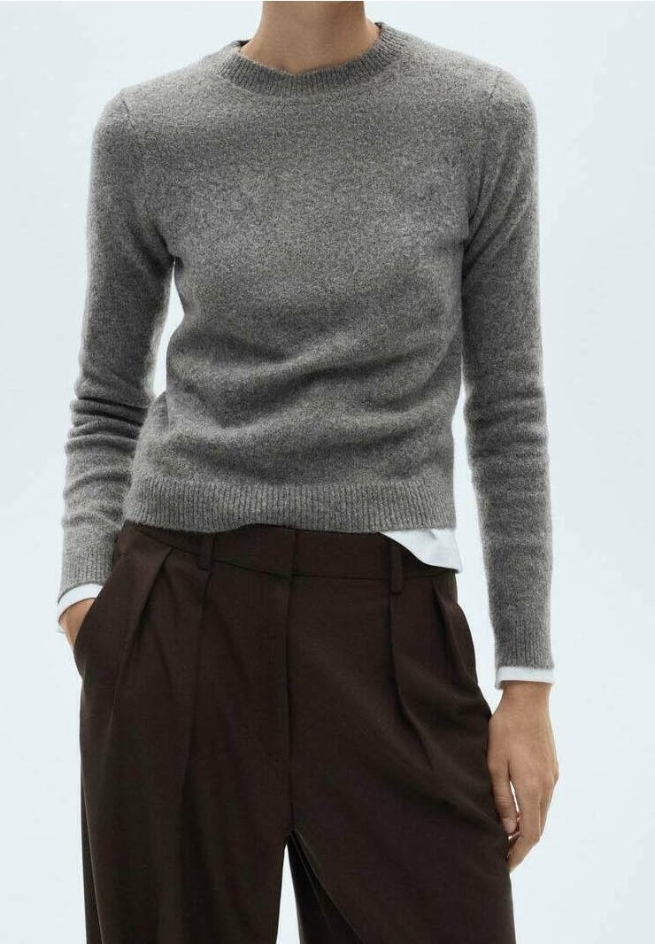 Personne portant un pull en laine gris ajusté sur une chemise blanche, associé à un pantalon plissé marron foncé à taille haute, mains dans les poches.