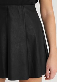 Jupe en simili cuir noire à design évasé, avec des plis et une texture lisse, main visible sur la taille.