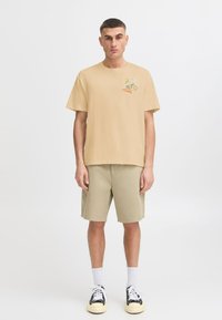 Light beige cotton t-shirt with a relaxed fit, featuring a graphic design on the left chest. Paired with light khaki shorts and black sneakers.