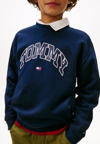 Marineblå sweatshirt med "TOMMY" i hvite bokstaver, med en liten rød, hvit og blå logo. Ribbestrikkede mansjetter og et lagdelt rødt helm nederst.