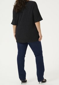 Femme se tenant dos à l'objectif, portant un haut noir texturé à manches courtes, un jean bleu foncé et des chaussures à talons noires sur fond blanc.