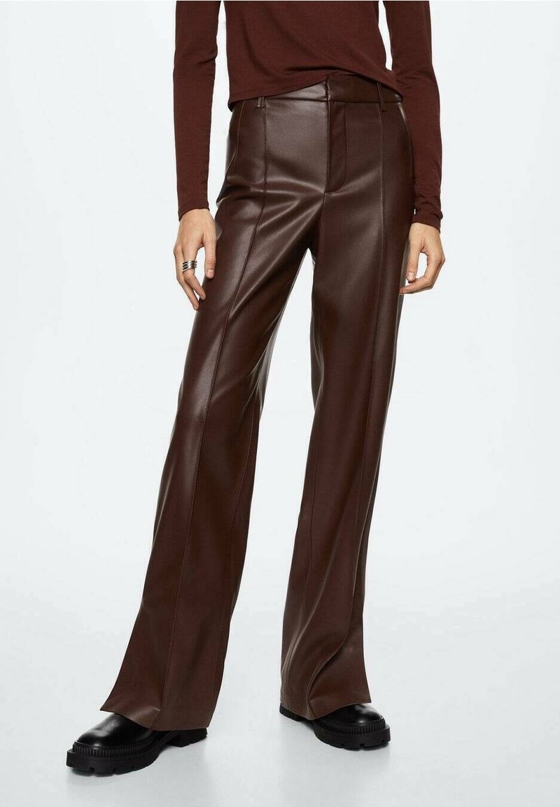 Femme portant un pantalon large en simili cuir marron foncé, associé à un haut marron à manches longues et des chaussures noires épaisses, debout sur fond blanc.
