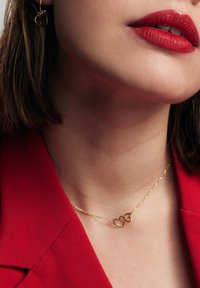 Złoty naszyjnik z dwoma splecionymi sercami na delikatnym łańcuszku; noszony z czerwonym żakietem. Gładka tekstura, minimalistyczny design.