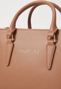 Tan syntetická kabelka s texturovaným povrchem, dvojitými horními madly, zlatými kovovými detaily a embosovaným logem 'VALENTINO' na přední straně.
