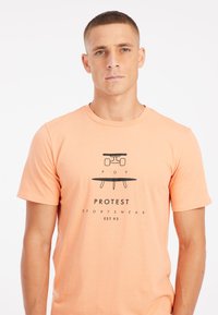 Perzikkleurig T-shirt met korte mouwen van katoen, met zwarte grafische print van een surfplank en de logo tekst "POV PROTEST SPORTSWEAR EST.93."