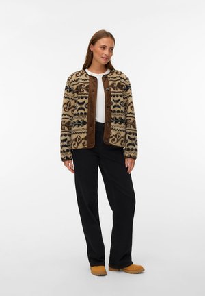 Mujer que lleva una chaqueta marrón y beige con botones y estampado, camisa blanca, pantalones negros y botas marrones claras, de pie sobre un fondo blanco.