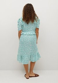 Vestido floral azul claro con mangas cortas con volantes, cintura peplum y dobladillo en capas. Lleva sandalias planas marrones. Se muestra la vista posterior.