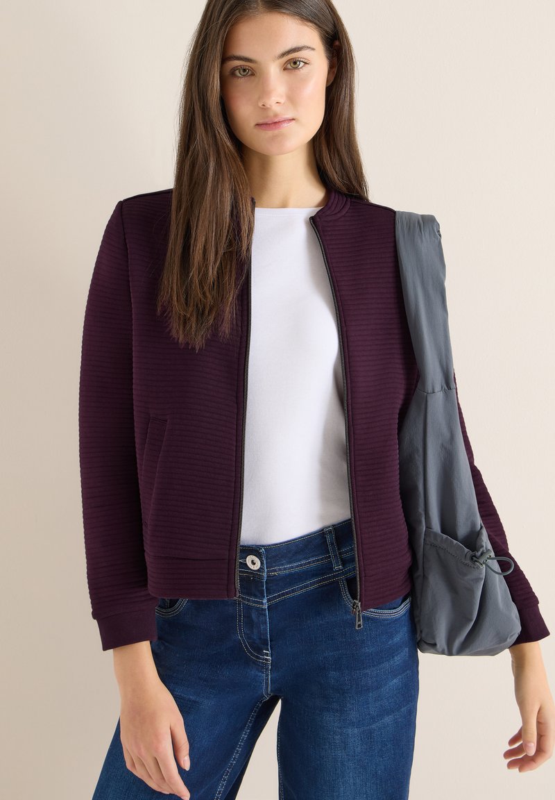 Bordeauxrode bomberjas met ribstructuur en een grijze schoudertas, gecombineerd met een wit shirt en blauwe jeans. De jas heeft een ritssluiting en zakken.