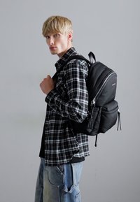 Rucsac negru, matlasat, purtat peste o cămașă din flanel cu un model în carouri alb-negru; are închidere cu fermoar și multiple compartimente.