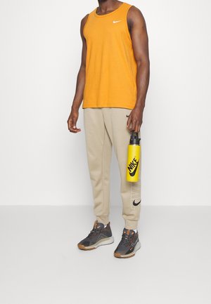 Mann trägt orangefarbenes ärmelloses Shirt und beige Nike-Sweatpants, hält eine gelbe Nike-Wasserflasche und steht vor weißem Hintergrund.