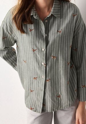 Kobieta nosząca zieloną koszulę zapinaną na guziki z białymi pionowymi paskami i małymi, haftowanymi wzorami brązowego psa i ptaka.