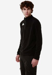 Pullover in pile nero con zip a metà, colletto alto e logo bianco sul lato sinistro del petto, abbinato a pantaloni scuri con tasche con zip.