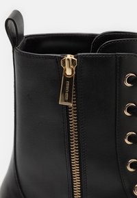 Stivale alla caviglia in pelle nera con zip dorata, occhielli in metallo e design con lacci. Texture liscia con tallone rinforzato.