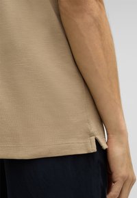 Beige strukturierte Bluse mit seitlichen Schlitzen, versehen mit einem dezenten Logo nahe dem Saum. Kombiniert mit dunkelblauen Hosen, die eine neutrale Farbpalette präsentieren.