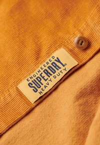 Orange corduroy-stof med et fremtrædende gult mærke, der står "ENGINEERED SUPERDRY HEAVY DUTY." Har en knapdetalje.