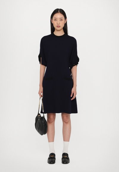 Robe marine à manches courtes avec deux poches avant noires, coupe décontractée, texture douce, associée à des mocassins noirs et un sac à main noir. Chaussettes blanches.
