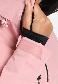 Hand, die den Reißverschluss einer rosa Jacke anpasst, wobei der schwarze Innenärmel mit Daumenloch und der rosa verstellbare Handgelenkriemen mit Klettverschluss zu sehen sind.