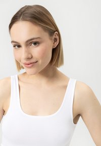 Jeune femme aux cheveux blonds courts portant un débardeur blanc, souriant légèrement, sur un fond blanc uni.