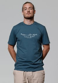 Man met teruggebonden haar en baard draagt een teal shirt met de Duitse tekst "Wenn nicht jetzt, wann dann?" en lichtbeige broek tegen een grijze achtergrond.