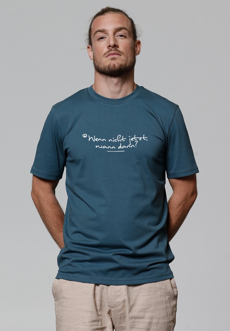 Man met teruggebonden haar en baard draagt een teal shirt met de Duitse tekst "Wenn nicht jetzt, wann dann?" en lichtbeige broek tegen een grijze achtergrond.