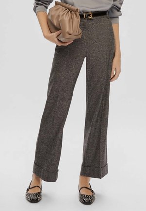 Donna che indossa pantaloni larghi grigi, cintura nera, ballerine nere borchiate, mentre tiene una borsa a mano beige a ruches su uno sfondo semplice.