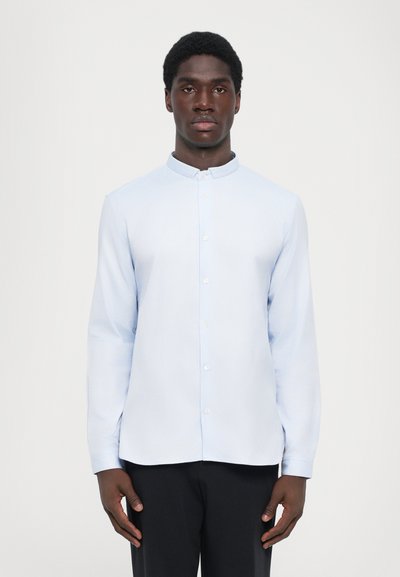 Chemise bleu clair à manches longues avec col mandarin, devant à boutons et rayures verticales subtiles. Texture lisse, coupe ajustée, design minimal.
