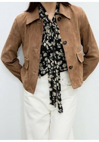 Veste en daim marron avec un col pointu, fermeture par boutons et poches avant, portée sur un chemisier noir à motifs floraux et un pantalon blanc.