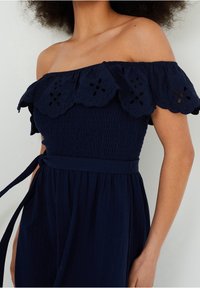 Navy off-shoulder jumpsuit met een gerimpelde, bloemenborduursel halslijn, smock lijfje en bijpassende tailleband. Zachte, gestructureerde stof.