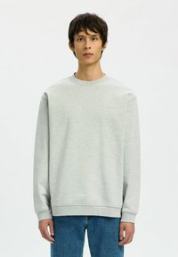Jeune homme debout, portant un sweat-shirt gris clair à col rond et un jean bleu, sur un fond blanc uni.