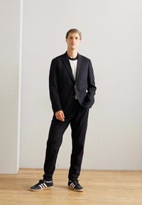 Mand står indendørs iført sort jakkesæt, hvid t-shirt og sorte sneakers med hvide striber, mod en simpel hvid væg og et trægulv.