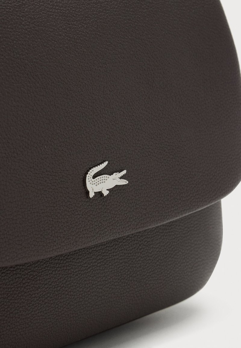 Primo piano di una borsa in pelle scura strutturata con piccolo emblema argentato del logo coccodrillo sulla patta frontale.