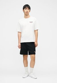 Άνδρας που φοράει λευκό μπλουζάκι Polo Sport, μαύρη βερμούδα, λευκές κάλτσες και λευκά αθλητικά παπούτσια, στέκεται μπροστά σε απλό φόντο.