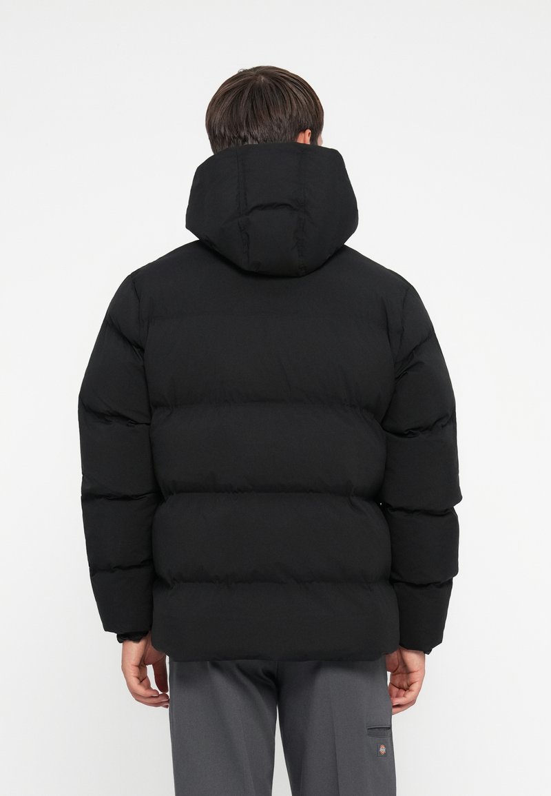 Chaqueta de plumas negra con capucha, con un diseño acolchado y un grosor notable. Construcción simple, costuras visibles y una silueta cuadrada.