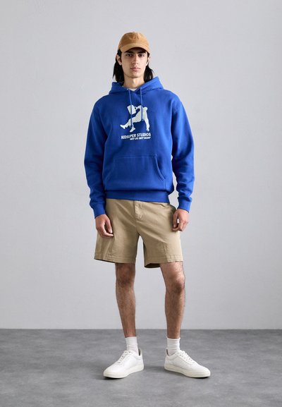 Felpa blu con stampa grafica bianca, pantaloni corti khaki e sneakers bianche. Uomo in piedi contro uno sfondo neutro con una posa informale.