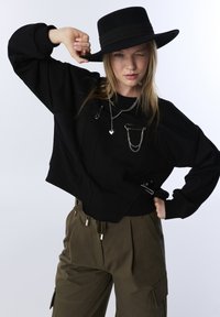 Zwarte cropped sweatshirt met ritsen en veiligheidsspelden, gedragen met olijfgroene broeken met plooien en zijzakken, op een witte achtergrond.