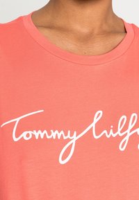 Zbliżenie na osobę ubraną w różową koszulkę z białym, kursywnym logo "Tommy Hilfiger" na piersi.