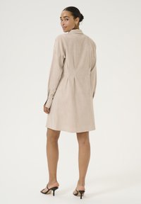 Robe beige, longueur genoux, en tissu doux, avec un col et des plis au dos. Associée à des sandales à talons noires.