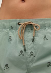 Shorts de bain vert clair avec un motif de crânes et de tibias noirs, taille élastique et cordon de serrage beige avec des extrémités noires. Texture lisse.