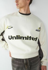 Kremowa bluza z nadrukiem "Unlimited" w wyraźnym czarnym kolorze. Posiada kontrastujące ciemne akcenty oraz małe logo na klatce piersiowej i rękawie.