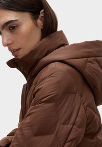 Brun, kviltet pufferjakke med høj krave og fastgjort hætte. Har hexagonal mønster og glat tekstur. Lynlås lukning foran.