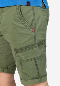 Khaki Cargo-Shorts mit strukturierter Oberfläche, seitlichen Taschen, umgeschlagenem Saum und einem kleinen roten Logo-Akzent in der Nähe der vorderen Tasche.