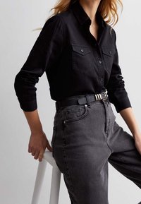 Donna che indossa una camicia nera con bottoni infilata in jeans grigi scuri a vita alta con una cintura nera, appoggiata a uno sgabello bianco.