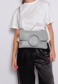 Borsa a mano in pelle grigia con un accento circolare, forma piatta, tracolla regolabile e finitura testurizzata, tenuta da una persona che indossa una camicia bianca.