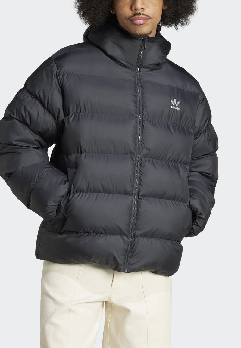 Schwarze Pufferjacke mit horizontalen Nähten, hohem Kragen, Reißverschluss und besticktem Logo. Über hellfarbigen Hosen getragen.