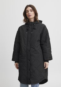 Sort quiltet lang jakke med hætte, der har trykknapper, snor og brede ærmer. Materialet ser glat ud med en mat finish.