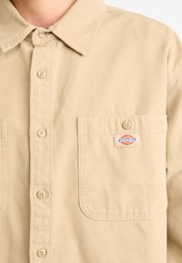 Camisa de botones beige hecha de tela de algodón, con cuello, bolsillo frontal y un pequeño parche con logo en el pecho.