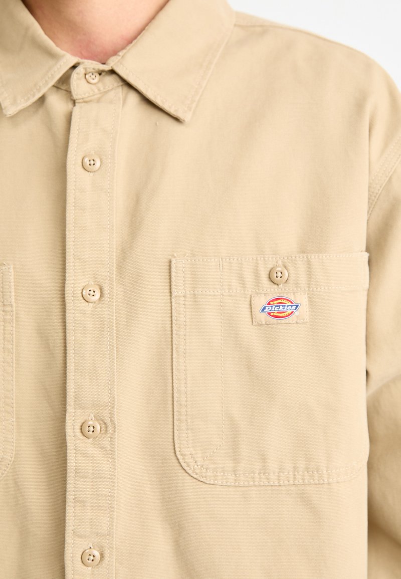 Camicia beige con bottoni realizzata in tessuto di cotone, dotata di colletto, taschino frontale e un piccolo patch con logo sul petto.