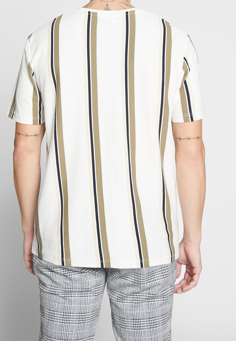 Homme portant une chemise blanche à manches courtes avec des rayures verticales beige et noires, associée à un pantalon gris à carreaux, vu de dos.