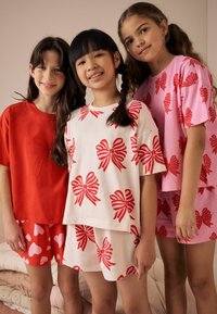 Tre ragazze indossano pigiami coordinati. Gli abiti presentano schemi di colori rosa e rossi con motivi a fiocco e cuori.